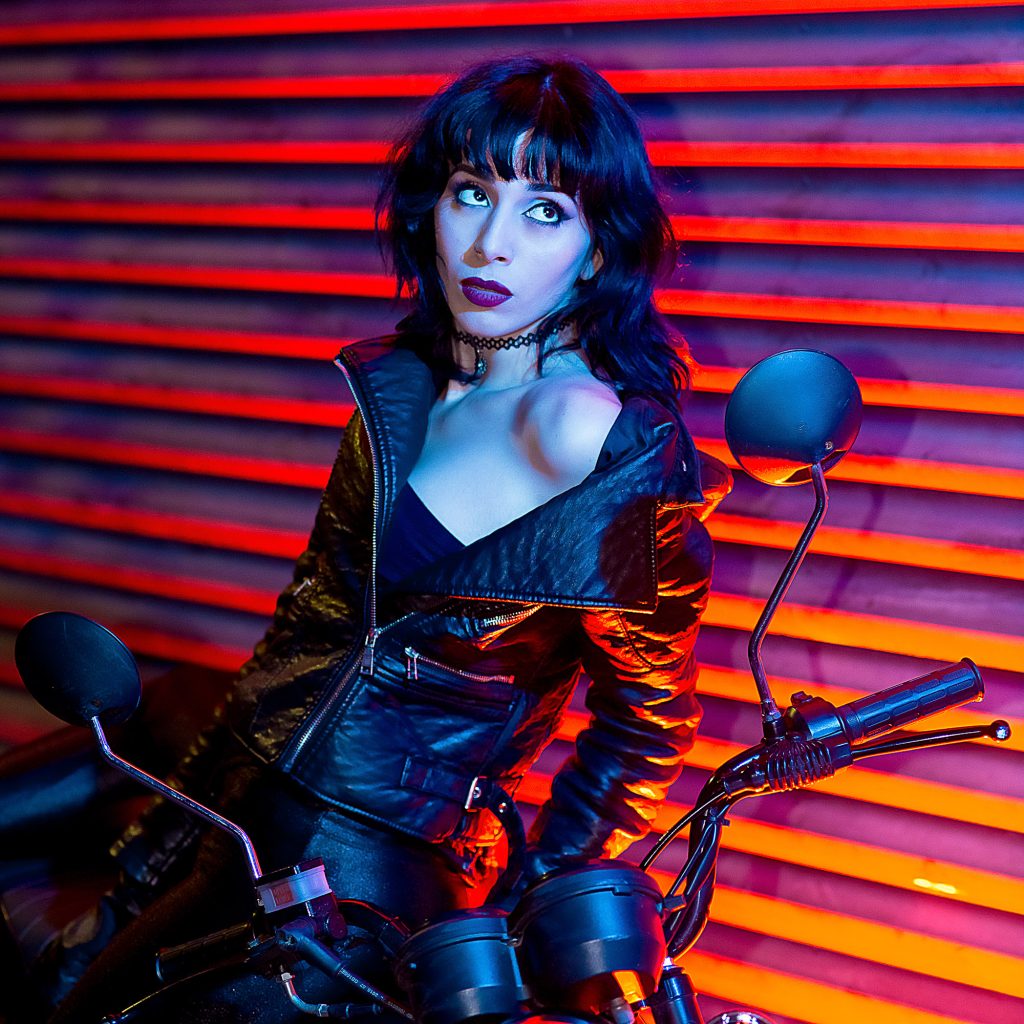 Retrato de Carmin chica motociclista con su moto. Iluminación strobist, softbox, mas rebote suave y fondo iluminado con flash rojo y azul contaminando la escena. Fotografía de retrato.