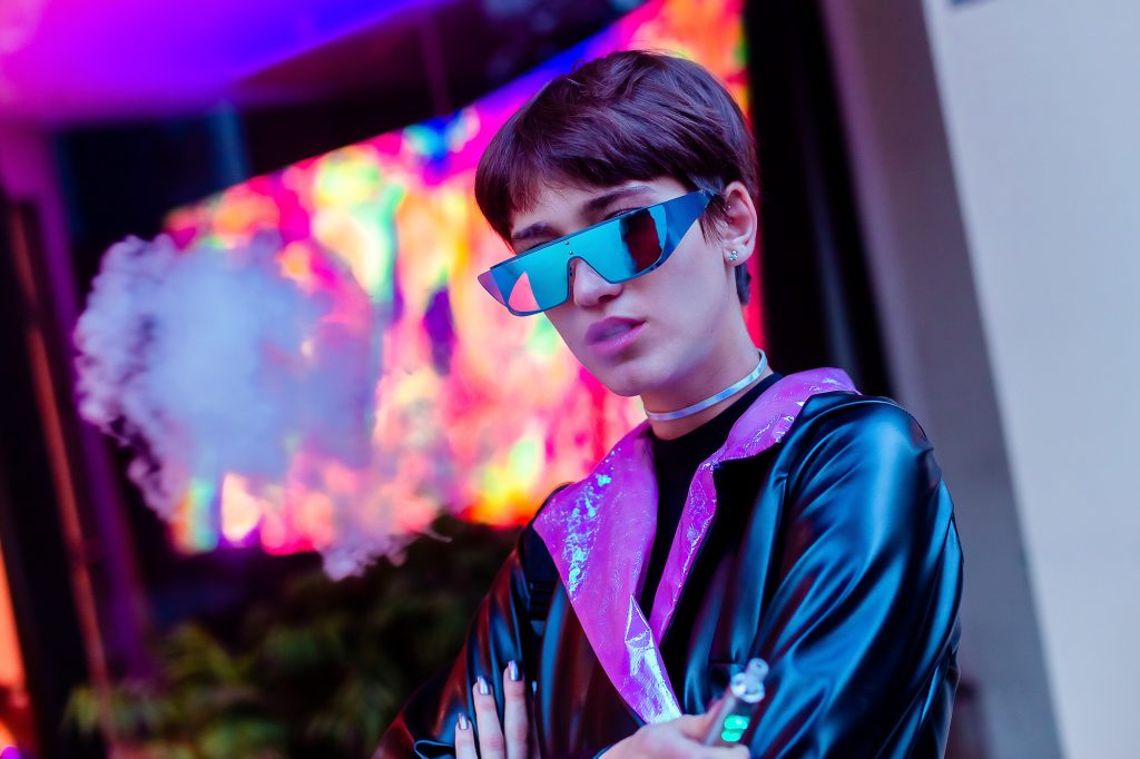 Modelo femenina en exteriores, look negro con rosa y negro iridiscente, lentes azules espejados y mochila holográfica.  Xona Retiro. Utiliza un vape como accesorio. Foto editorial de estilismo propio.