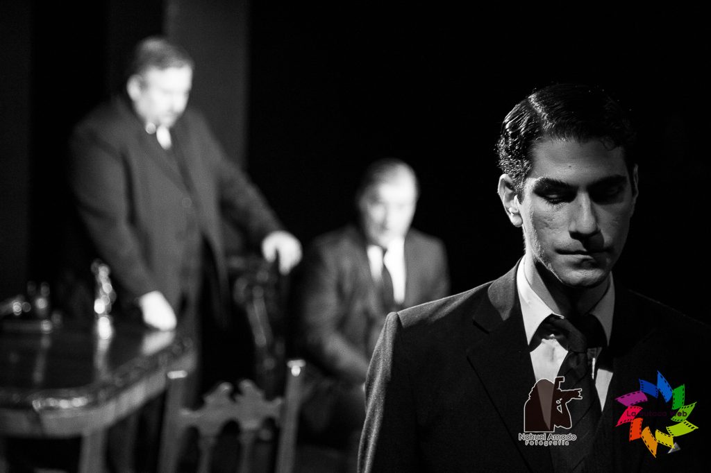 Foto de la obra teatral hombres de honor para la butaca web B&N hombre al frente en penumbra con mirada sospechosa y 2 al fondo iluminados fuera de foco
